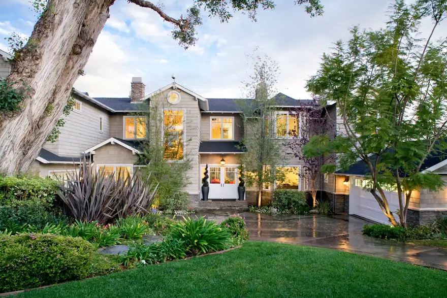 Shingled two story home at dusk with lit windows and a landscaped front garden